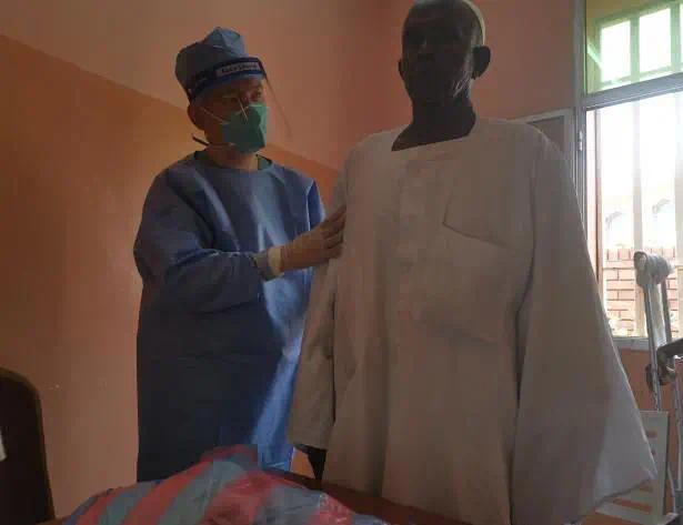 图为：王国军医生为苏丹腰痛患者治疗前患者只能弯腰在他人搀扶下行走，治疗后即可伸直腰独自行走